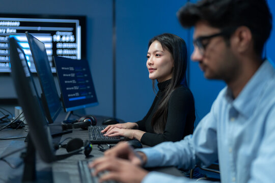 South Asian man and Asian woman code side by side at adjacent monitors filled with code in a dark blue software development office at night.