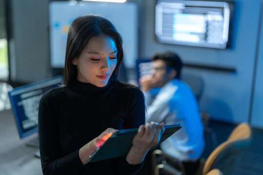 Young Asian female IT specialist working late on a digital tablet in a dark, blue-lit modern office, with a colleague working in the background.