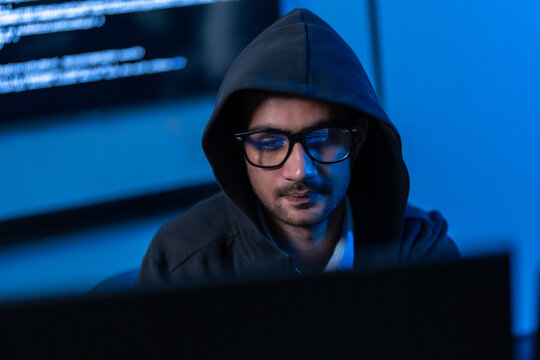 South Asian man in a hood and glasses focuses on a screen of code in a dark blue room, evoking cybersecurity, hacking and online threat themes.