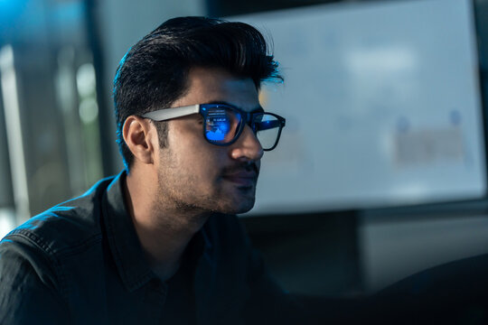 Side profile close-up of a South Asian man in glasses, a screen of data reflected on his lenses as cool blue light falls across his face at night.