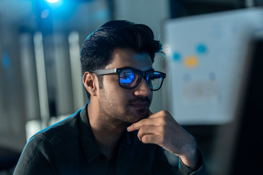Close-up of a South Asian man in glasses resting his hand on his chin in thought, a screen reflected on his lenses in a dark blue office.
