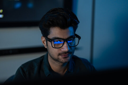 Tight portrait of a South Asian man in glasses looking down in concentration, a faint screen reflection on his lenses in a dark blue room.