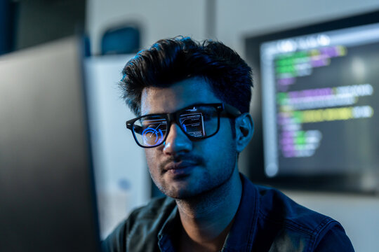 Close-up of a South Asian man in glasses with colorful code reflected on his lenses as he focuses on a screen in a dark blue tech office.