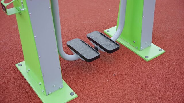 Elliptical trainer with black foot pedals and sturdy metal frame. Machine securely bolted to red rubberized safety surface in sports park