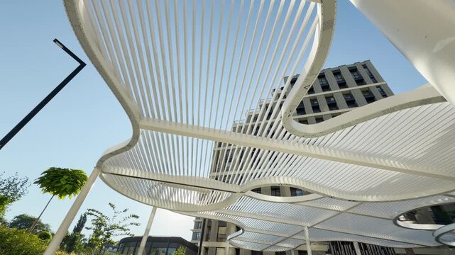 A curved white canopy stretches beautifully across a sunny courtyard in Simferopol, Crimea. Sunlight filters through the modern design, evoking a peaceful and cheerful feeling.