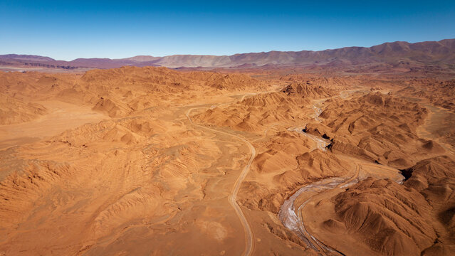 Aerial view of the arid desert landscape with winding dirt roads and dry riverbeds surrounded by rolling hills under a clear blue sky in Tolar Grande, Salta Province, Argentina.