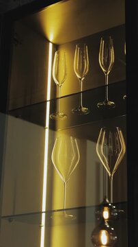 Vertical video. Wine glasses on illuminated glass shelves inside a modern display cabinet.