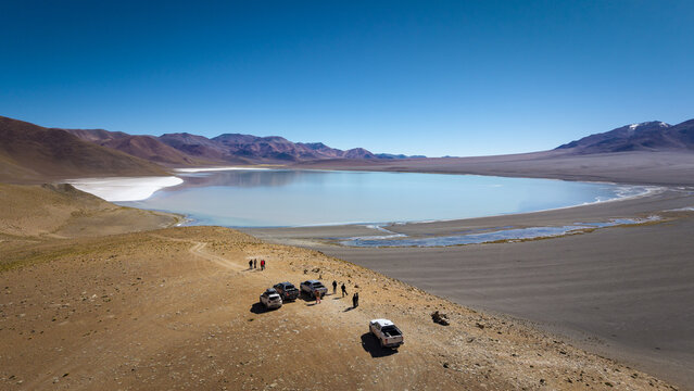 Aerial view of the landscape near El Penon, featuring a serene lake, arid mountains, off-road vehicles, and people in El Penon, Catamarca Province, Argentina.