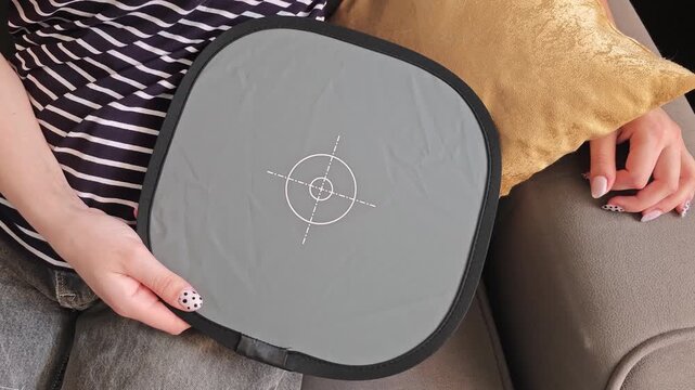 Female model in casual attire holds a gray reflector disc with target markings while seated on a couch, showcasing the items details and texture in a cozy indoor setting