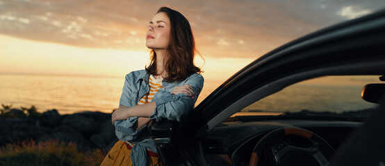 Fototapeta premium A reflective woman leans against a car at sunset on a coastal road, wearing a denim jacket and a yellow bag. A calm mood for lifestyle and travel storytelling.