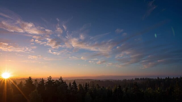 Sunrise above a forest landscape captured in timelapse. Fast moving clouds sweep across the sky while golden morning light illuminates the horizon and treetops below.