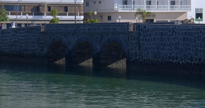 Wide shot of the stone bridge at Calle Punta de la Lagarta, with Arrecife in background