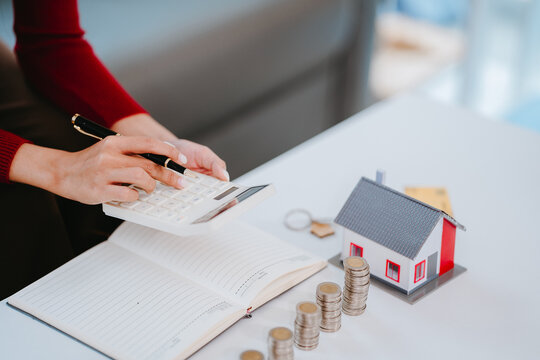Cropped shot of a woman writing in notebook with stacked coins, miniature house, and blue piggy bank on table, property investment and financial planning concept.