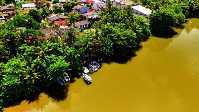 Small motorboats sit along a dense tree-lined Nilwala River bank beside the residential Matara neighborhood on a bright day.