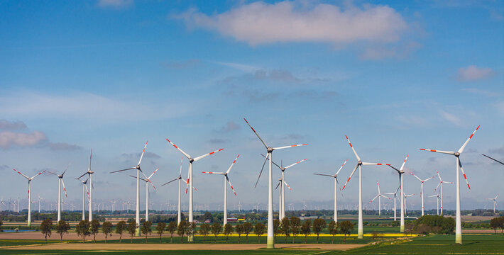 Aerial view of rows of wind turbines standing in a green agricultural field under a blue sky in Germany.