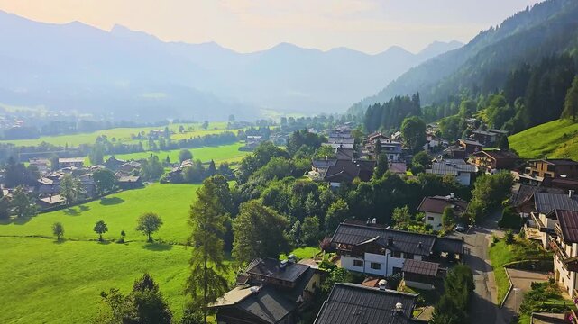 Elevated cable car view moving across a green alpine valley, with village homes, rolling fields, dark forest, and layered blue mountains under strong afternoon sunlight.