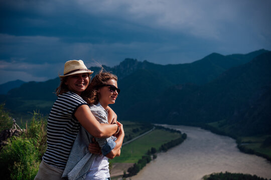 Two female travelers embracing on a mountain viewpoint above a winding river valley, enjoying a scenic outdoor adventure under dramatic storm clouds. Lesbian couple's jorney