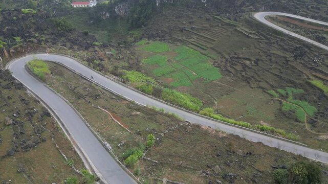 Top down aerial drone view of a motorcyclist riding along a winding mountain hairpin road through terraced farmland and rugged limestone terrain at Ma Pi Leng Pass in Ha Giang Vietnam