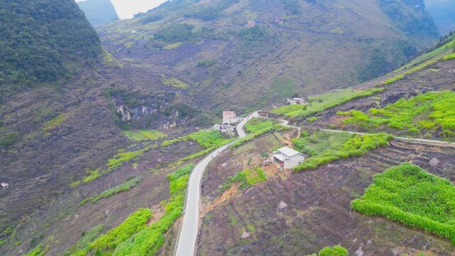 Aerial drone view of a winding road passing through terraced farmland and scattered rural homes, surrounded by rugged limestone terrain in Dong Van Karst Plateau Ha Giang Vietnam