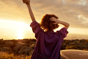 A woman with curly hair stands on a sandy beach at sunset, arms raised in joy. Warm light and open sky convey freedom, confidence, and a relaxed lifestyle moment. © SHOTPRIME STUDIO