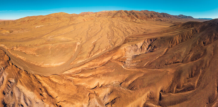 Aerial view of the arid, rugged desert landscape with deep erosion gullies, rolling hills, and vast sandy terrain under a clear blue sky in San Pedro de Atacama, Antofagasta, Chile.