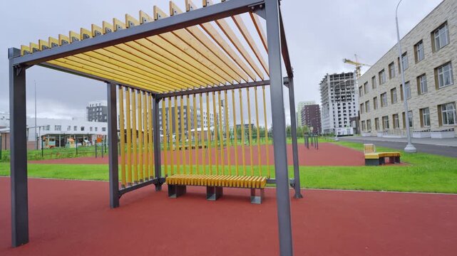 Wooden bench sits in modern slatted pergola on red sports ground. Contemporary architecture creates clean geometric lines in urban park