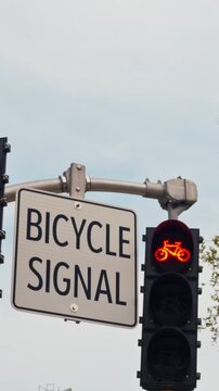 Bicycle signal sign beside traffic light glows red, mounted on street pole against sky, with illuminated bike icon indicating bike lane crossing control at intersection during stop phase.
