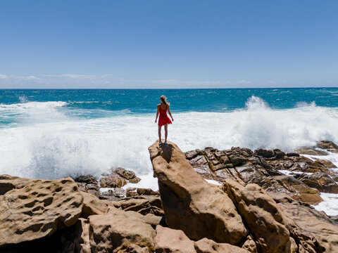 Aerial view of a woman in a red dress standing on a rock at Injidup Natural Spa with crashing waves and turquoise water in Yallingup, Western Australia, Australia.