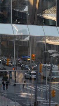 City traffic seen through glass building reflection with cars, pedestrians, crosswalk markings, and streetlights in urban downtown intersection as light shifts across mirrored facade