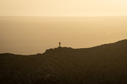 22 January 2022: View of the backlit silhouette of a person standing on a rocky mountain ridge overlooking a hazy valley under an orange sky during a golden sunset. Calderon Hondo