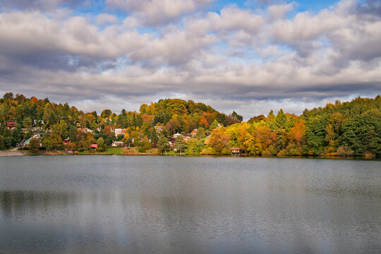 View of Velka Richnava lake with colorful autumn trees and hillside houses under a cloudy sky in Stiavnicke Bane, Banska Bystrica Regio, Slovakia.