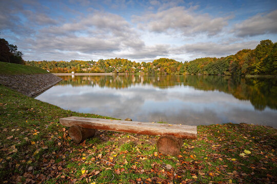 View of Velka Richnava lake with a wooden bench on the grassy shore surrounded by autumn trees under a cloudy sky, Stiavnicke Bane, Banska Bystrica Regio, Slovakia.