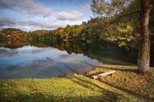 View of Velka Richnava Lake surrounded by colorful autumn trees and reflections in the water with a wooden bench in the foreground in Stiavnicke Bane, Banska Bystrica Region, Slovakia.