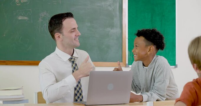 Male adult teacher uses laptop beside young boy student, guiding back to school elementary lesson, question practice, digital classwork, positive support and first day learning routine together