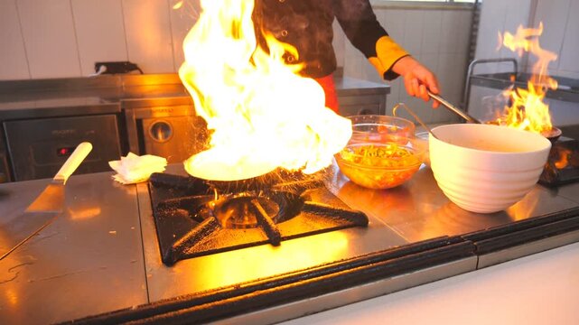 Unrecognizable chef holding two flaming pans and cooking tasty flambe style dish in modern kitchen of hotel. Professional cook working with different ingredients in cuisine. Dolly shot Slow motion