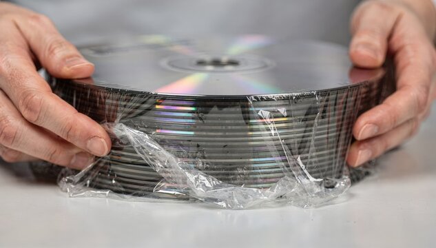 Hands carefully positioning shrinkwrap around replicated media discs emphasizing the shiny sealed surface with the background softly blurred for attention.