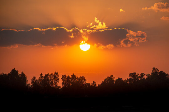 View of a vibrant sunset with the sun partially obscured by dark clouds above a silhouette of dense trees in Bogura, Rajshahi Division, Bangladesh.