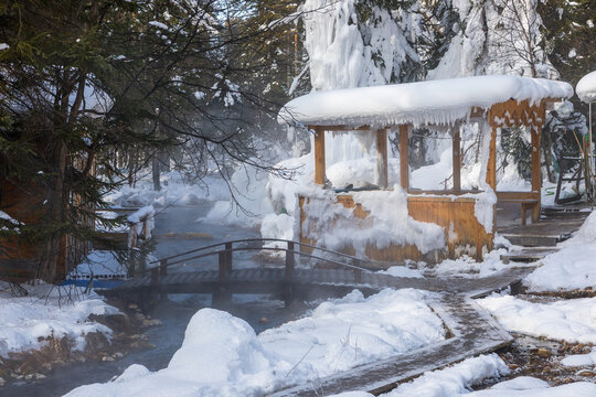 View of Goryachiye Istochniki Khakusy with a wooden bridge over a steaming thermal spring, snow-covered trees, and a wooden gazebo in Severobaykal'skiy Rayon, Respublika Buryatiya, Russia.