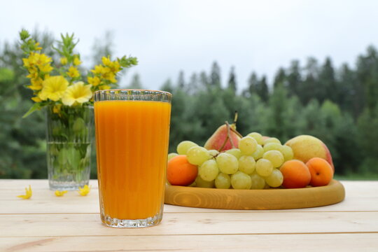 On a wooden desk, on the background of a summer forest landscape, there is a summer still life: a glass of orange juice, wooden tray of fruits and a glass jar of flowers. 
