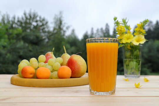 On a wooden desk, on the background of a summer forest landscape, there is a summer still life: a glass of orange juice, wooden tray of fruits and a glass jar of flowers. 