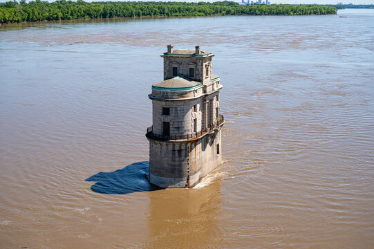 View of the historic water intake tower rising from the Mississippi River, with calm brown water and lush green trees along the distant riverbank in St. Louis, Missouri, United States.
