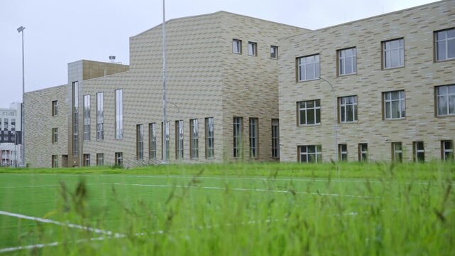 Large modern school building with tan brick facade. Professional green football field with markings visible behind patch of long grass