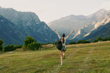 Obraz premium A woman with a backpack stands in a grassy meadow, arms raised in triumph, with expansive mountains behind. Concept: freedom, exploration, and active lifestyle.