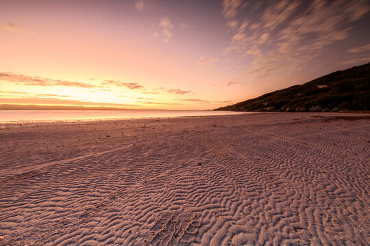 View of Quaranup Beach at sunset with rippled sand patterns on the shore and a calm sea under a soft orange sky in Vancouver Peninsula, Western Australia, Australia.