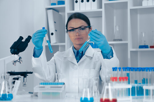In a wellequipped lab, a focused female scientist in protective gear examines two test tubes with different substances, surrounded by glassware and advanced equipment for her research
