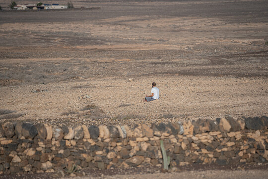 24 January 2022: View of a man seen from behind sitting in meditation in a vast arid rocky landscape with a stone wall and small cactus in the foreground.