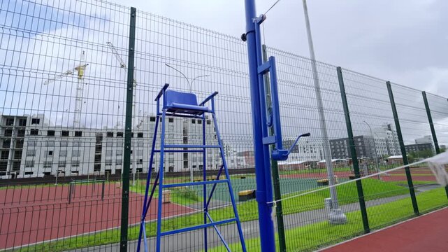 Referee chair stands by volleyball net on red sports ground. High-rise construction and cranes create industrial backdrop for stadium