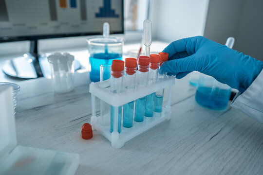 Close up of a gloved hand organizing test tubes with blue solution on a white laboratory table. Advanced scientific research and chemical analysis are taking place in a high tech lab