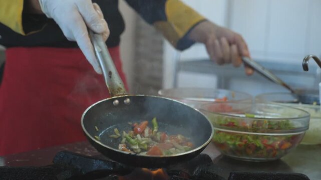 Unrecognizable chef holding two pans and cooking flambe style dish in modern kitchen of hotel. Male cook tossing and frying vegetables in flaming skillets on stove. Concept of cooking. Slow motion