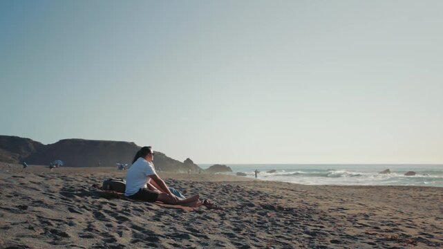 Couple sitting on the sand as the guy points at something in the ocean. Peaceful moment with sea horizon and rocky coastline in the background